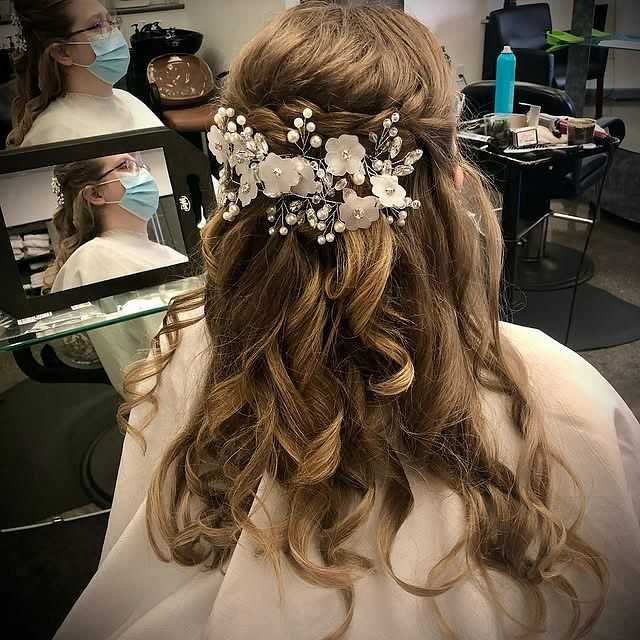 Bride with curled hair adorned with white floral hairpiece, looking in a salon mirror.