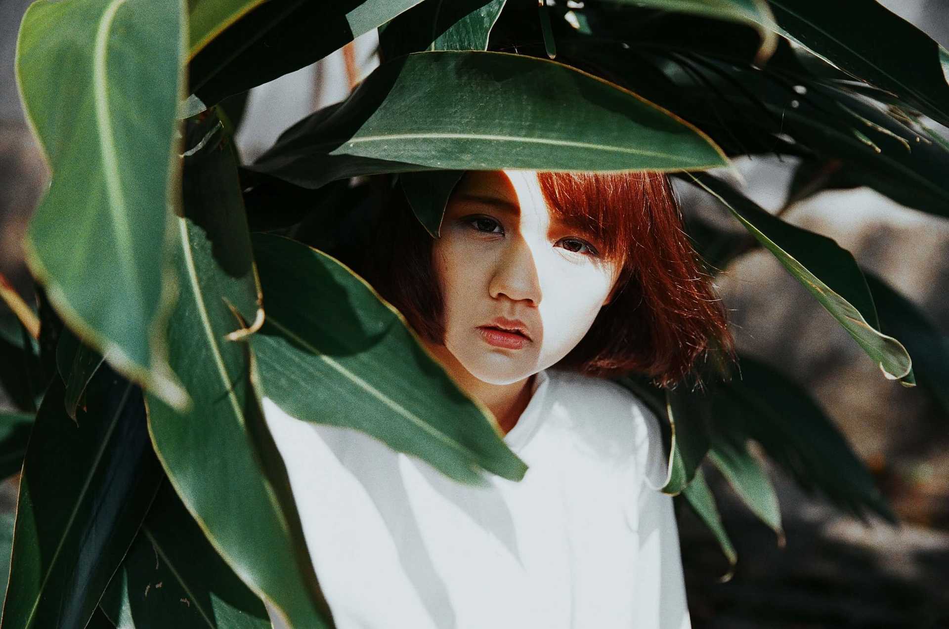 Person peeking through large green leaves, partially shaded, with a serious expression.