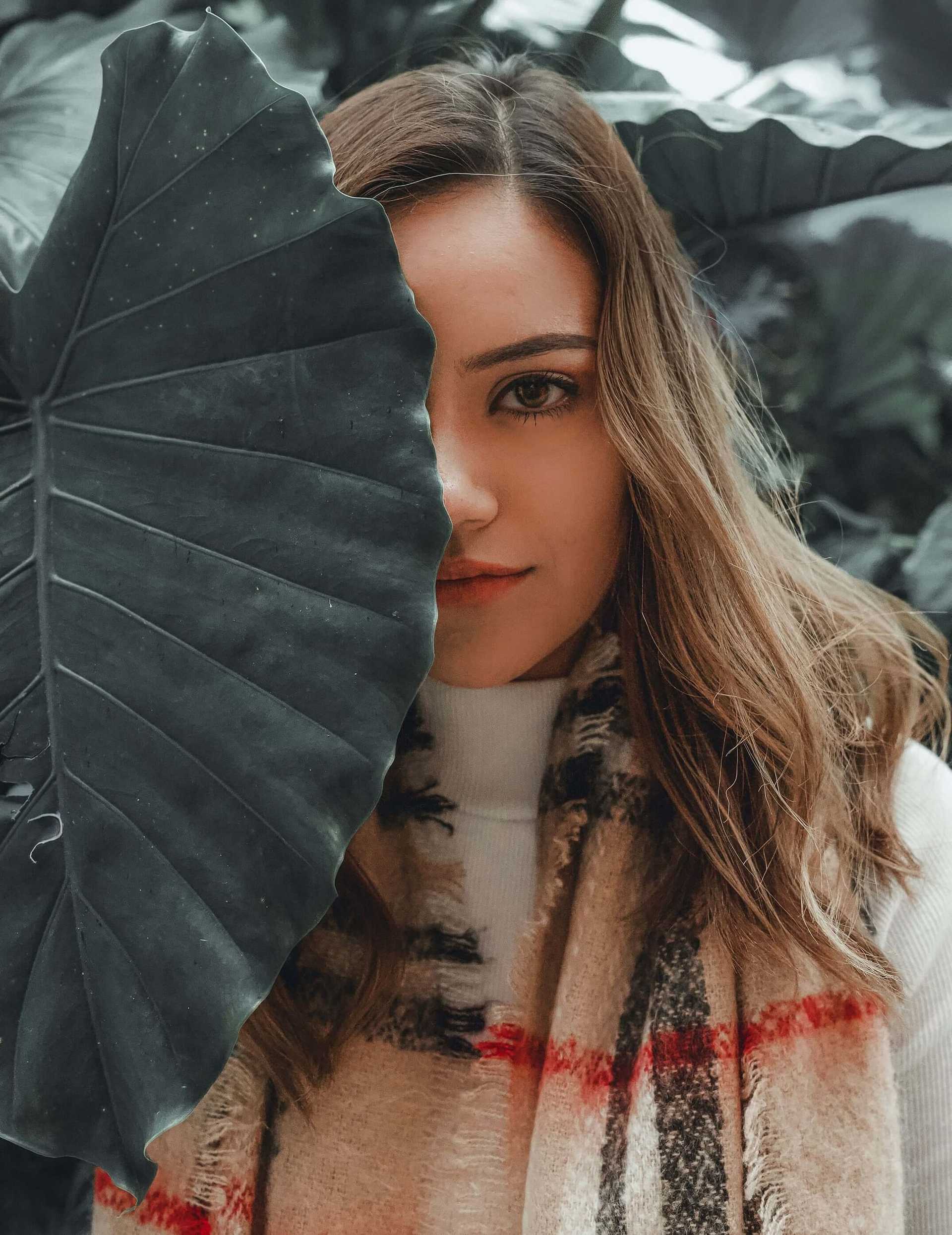 Woman partially hidden by large leaf, wearing a scarf, amidst lush greenery.