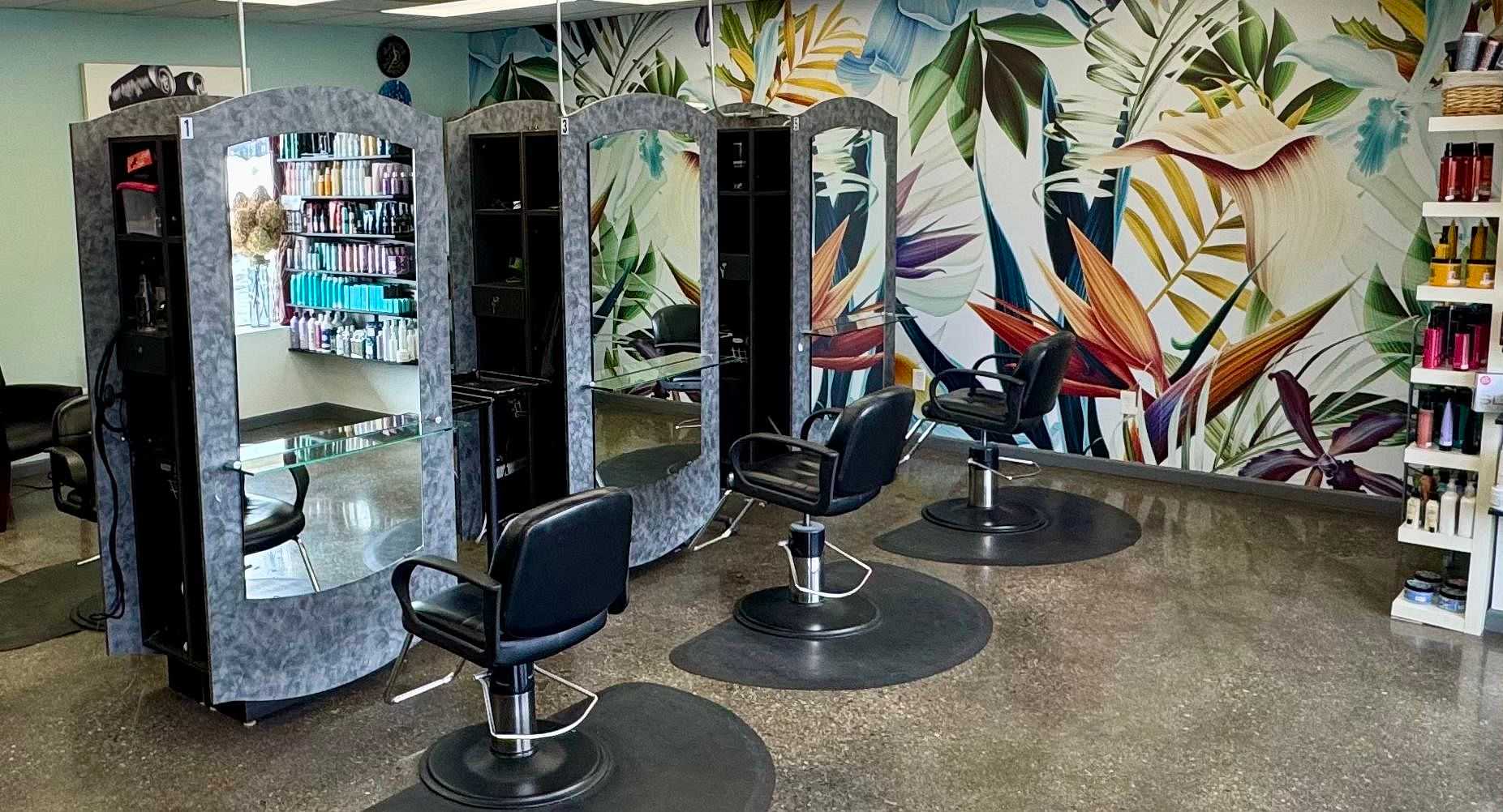 Modern hair salon with mirrors, tropical wall, styling chairs, and shelves of products.