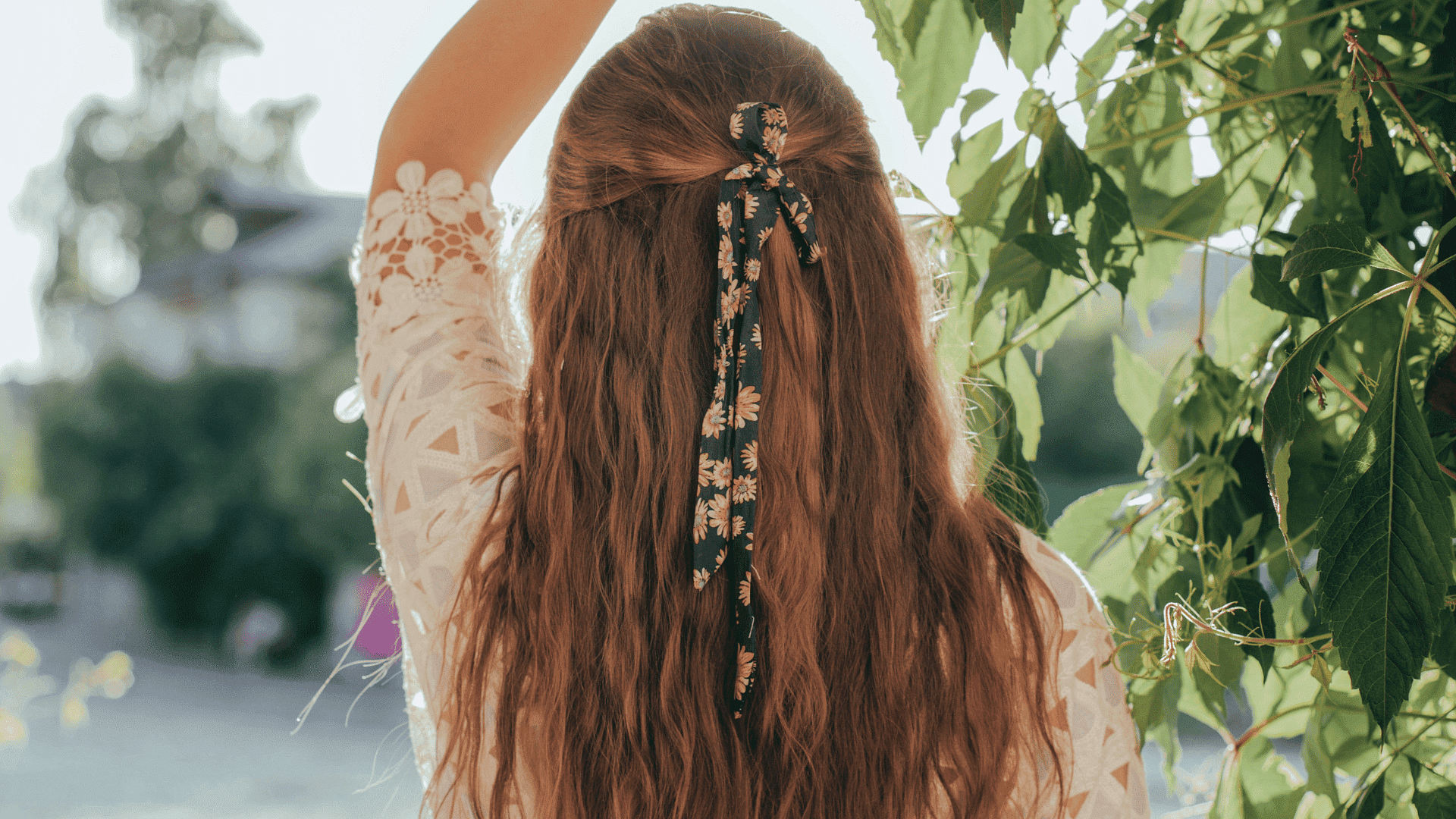 Woman with floral hair ribbon, standing outdoors, surrounded by green foliage.